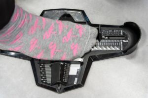 A foot wearing a gray sock with pink designs is placed on a shoe sizing device, offering a helpful guide for those with wide feet. The device measures the length and width, showing different size markings to ensure perfect footwear tips.